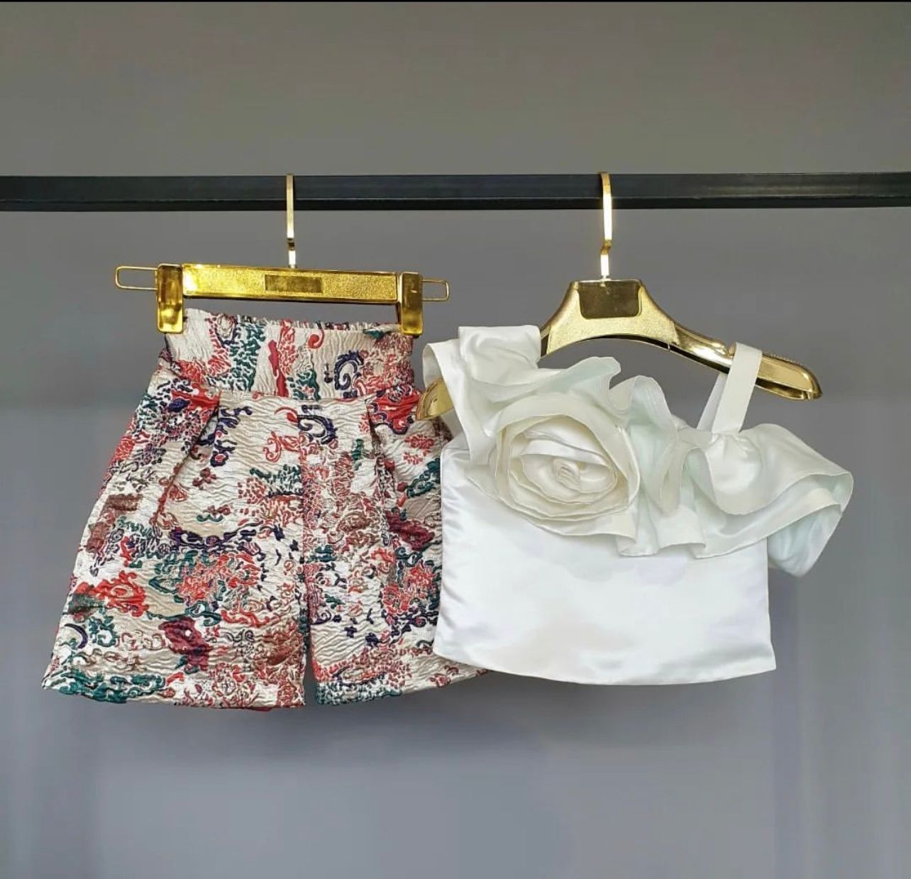 A gold-plated shorts with orange shimmery thread and a white satin top with a round silver-plated brooch on waist, displayed on a hanger.