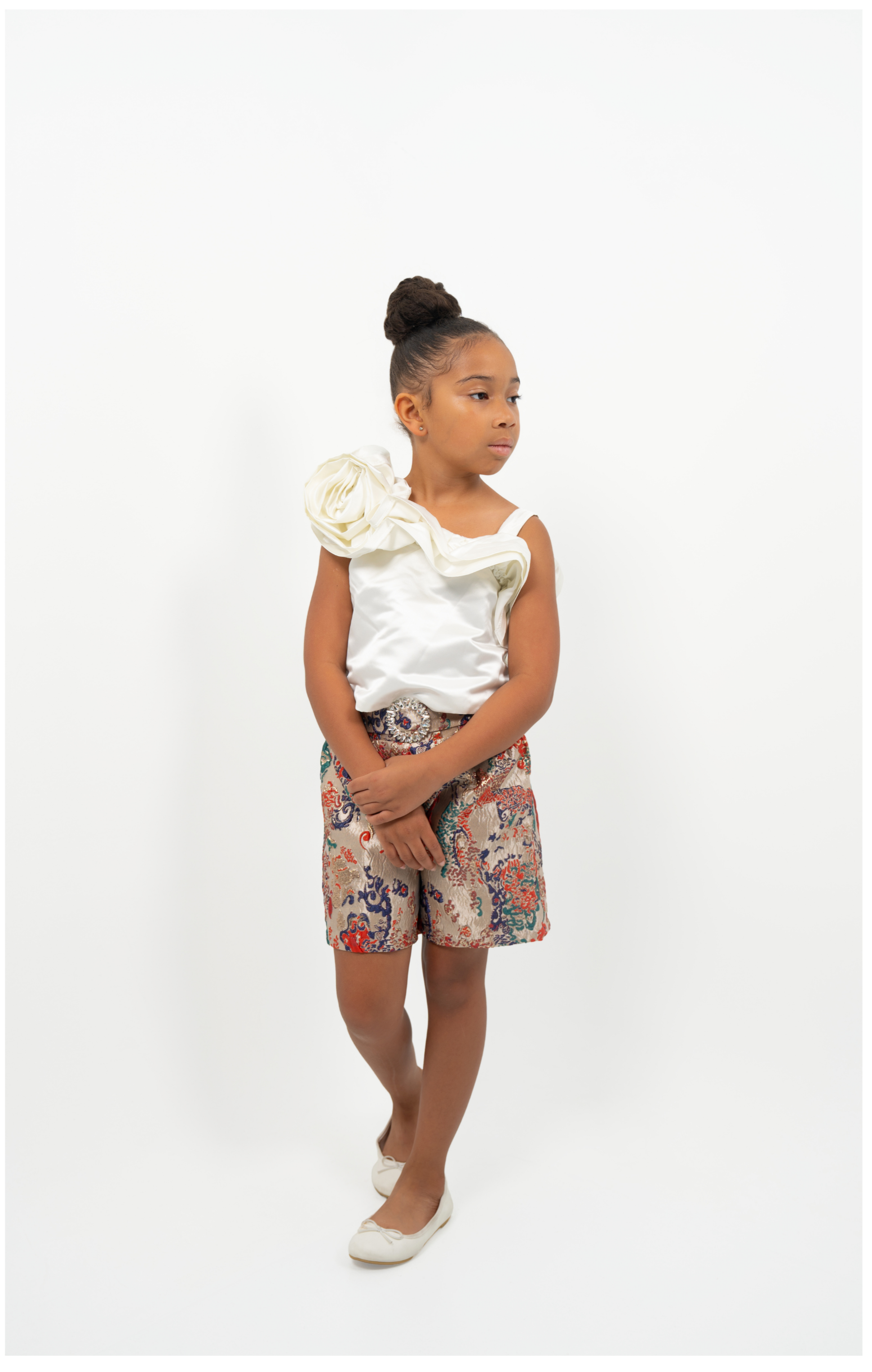 Young Girl wearing white satin flower-shoulder top with brocade shorts, styled for elegant party or special occasion

