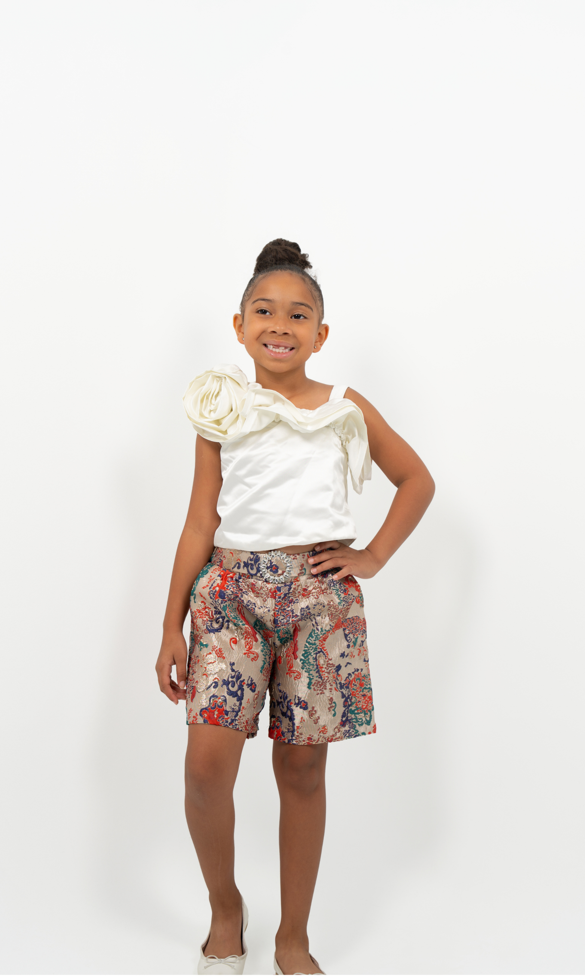 Young Girl wearing white satin flower-shoulder top with brocade shorts, styled for elegant party or special occasion
